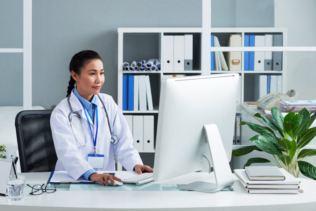 Asian female doctor in white lab coat working on computer for Lean Healthcare Overview Training, likely learning about basic principles and tools of Lean methodology to improve patient care and increase efficiency in healthcare.
