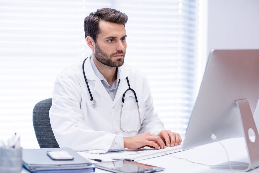 Male doctor in white lab coat working on computer for Lean Healthcare Overview Training, likely learning about basic principles and tools of Lean methodology to improve patient care and increase efficiency in healthcare.