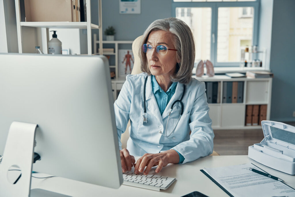 Serious mature female doctor in white lab coat working on computer for Lean Healthcare Specialist Training and Certification, likely using Lean methodologies to improve processes and provide better patient care.