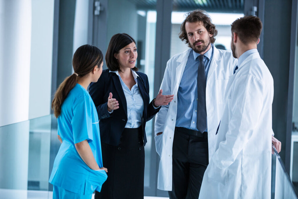 Group of healthcare providers and executive having a Lean meeting in hospital hallway, discussing ways to improve patient care and streamline processes, with a woman manager leading the discussion.