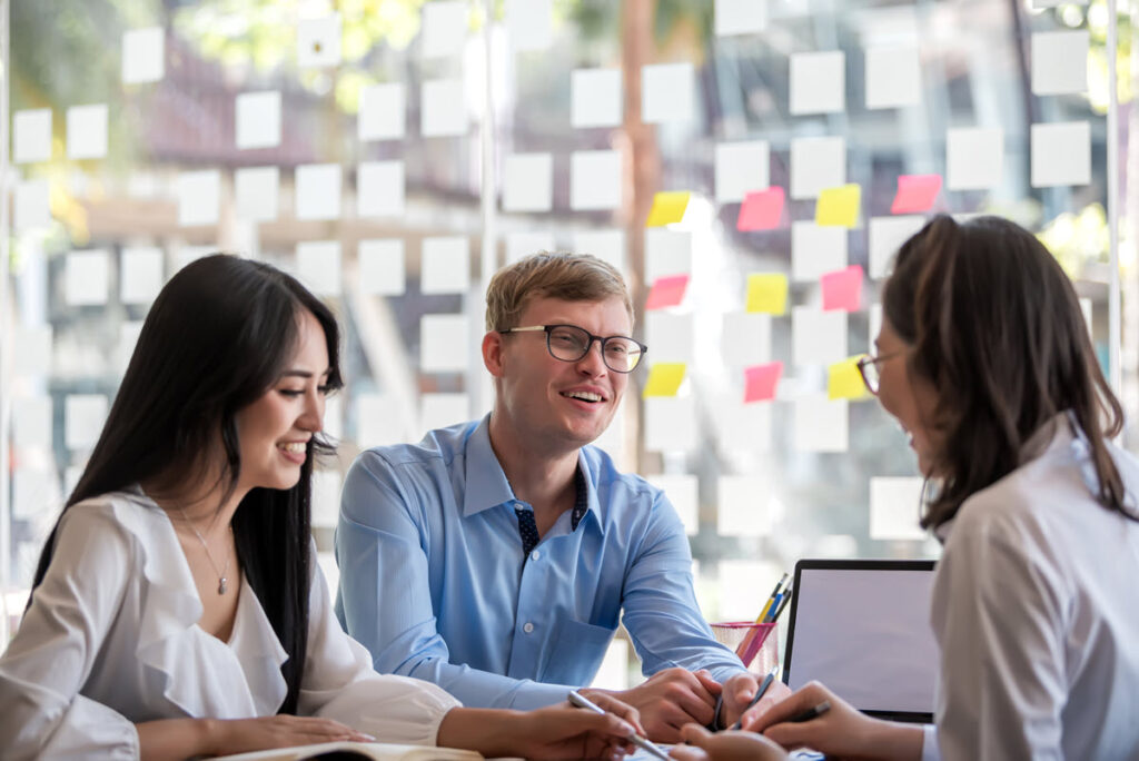 Group of modern, smartly dressed people meeting for Lean Six Sigma White Belt Training, collaborating and brainstorming solutions to improve processes, with a glass wall in the background displaying post-it notes for process mapping.