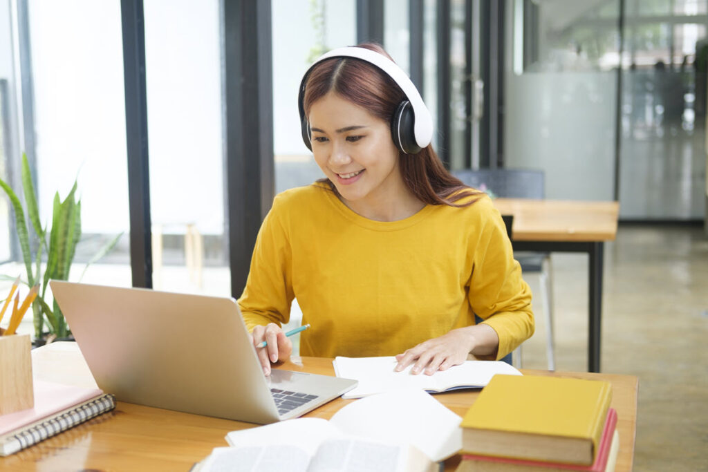 Asian woman wearing headphones working on Lean Specialist Training and Certification on her laptop, likely using data analysis, process mapping, or statistical tools to reduce waste in processes using Lean methodologies.